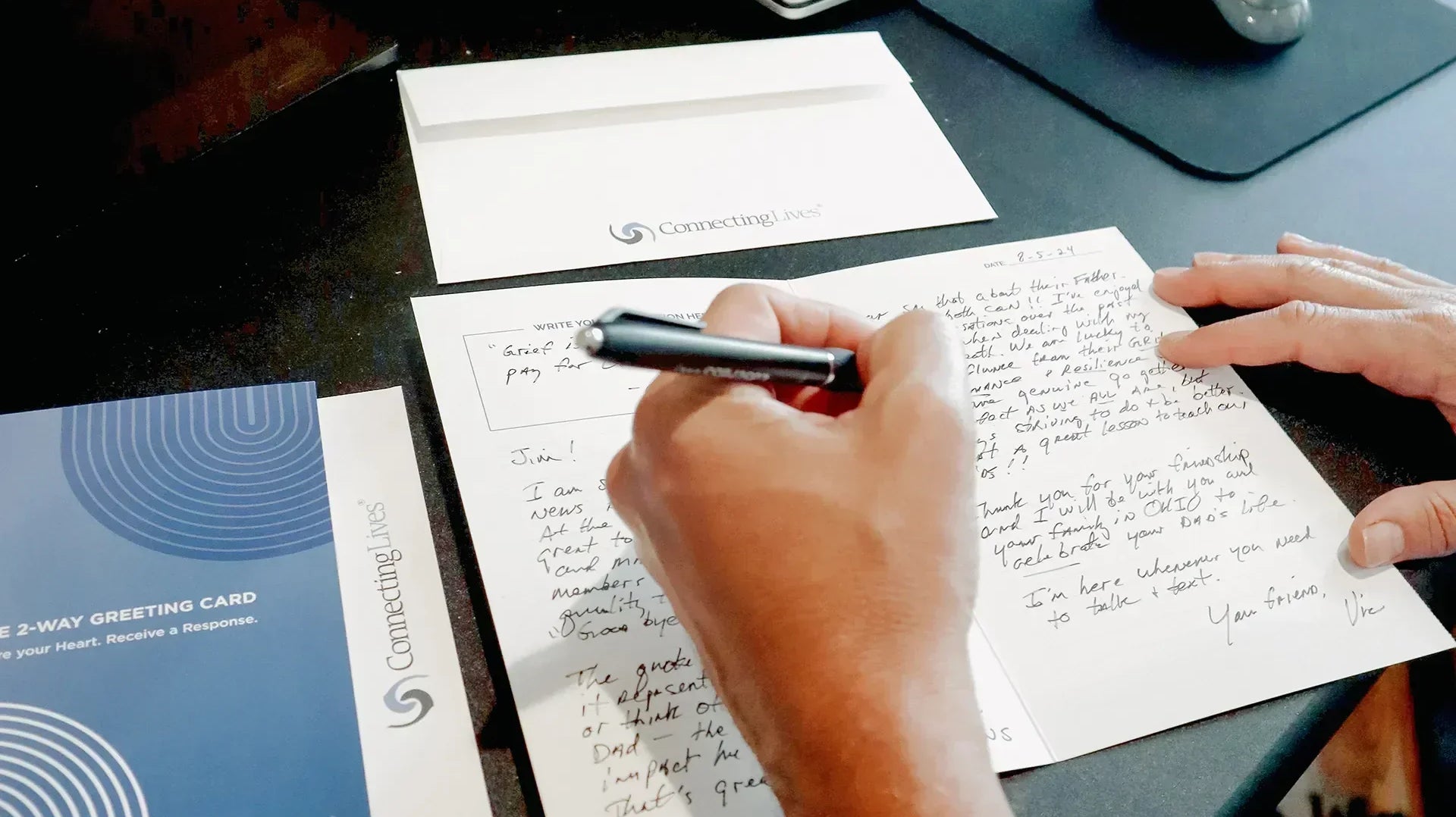 Person writing a heartfelt letter with a ConnectingLives 2-Way Greeting Card and envelope on a desk