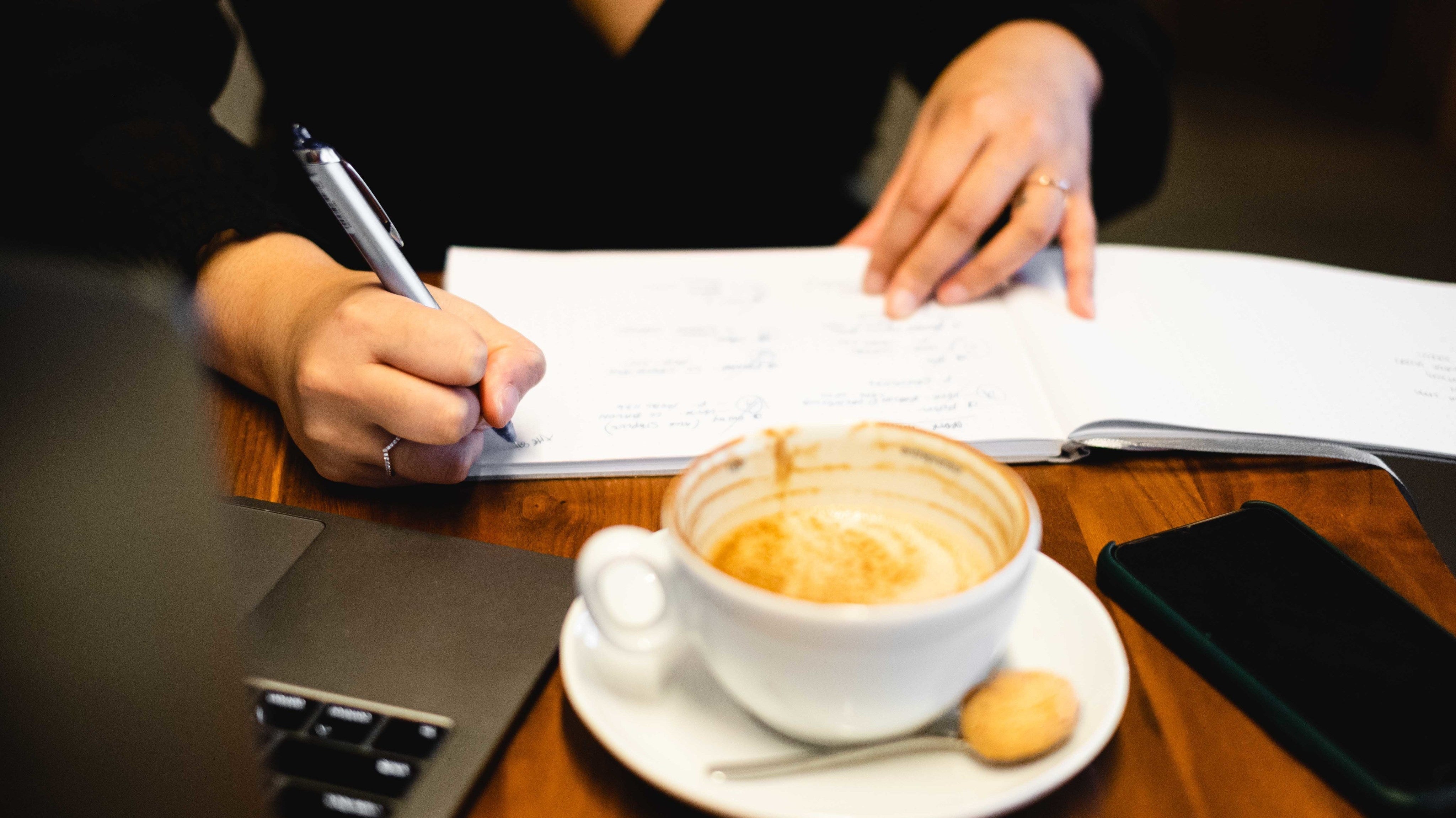 Hands writing in an open notebook next to a coffee cup — a moment of thoughtful connection