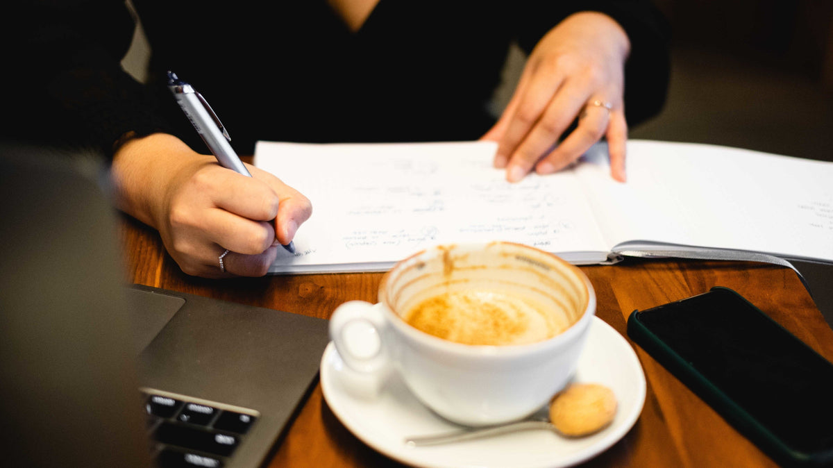 Hands writing in an open notebook next to a coffee cup — a moment of thoughtful connection